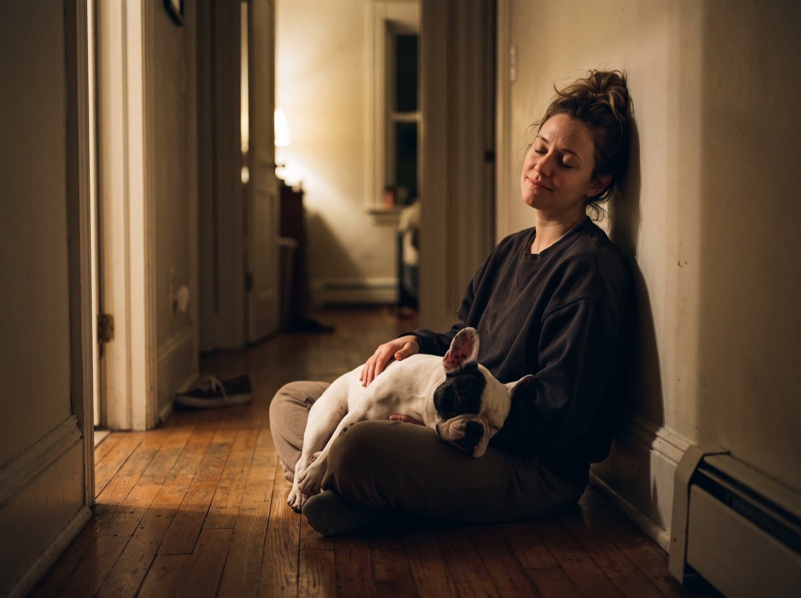 A tired woman sitting on a dark hallway floor with her French Bulldog asleep in her lap